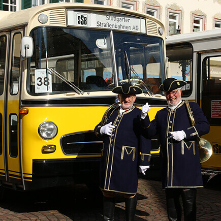 Gelber Oldtimerbus und zwei Personen in traditioneller Kleidung beim Oldtimer Bustreffen auf dem Marktplatz in Bad Mergentheim.